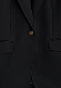 Blazer noir sur mesure avec un unique bouton doré orné au centre et deux poches à rabat devant de chaque côté.