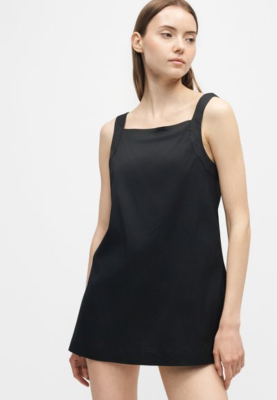 Jeune femme portant une mini-robe noire sans manches avec de larges bretelles, debout devant un fond clair uni, regardant sur le côté.