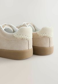Beige suede sneakers with a cream accent at the heel, featuring a stitched detail and a textured brown rubber sole.
