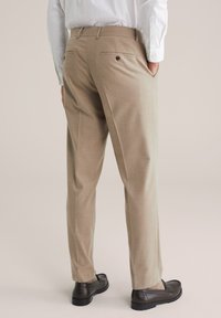 Homme se tenant avec les mains dans les poches, portant un pantalon habillé beige, une chemise blanche, des chaussettes beige à motifs et des mocassins en cuir marron foncé.