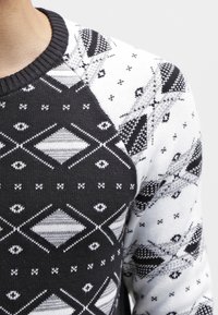 Pull tricoté présentant un motif géométrique noir et blanc, avec un col côtelé et des manches contrastantes, affichant des motifs de diamants et d'yeux.