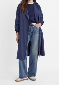 Marineblauwe trenchcoat met dubbele knopen en zijzakken, gedragen over een marineblauwe trui en lichtblauwe wijde broek, gecombineerd met sneakers.
