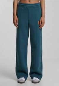 Pantalones de chándal de pierna ancha en color verde azulado, fabricados en tejido suave, con cinturilla elástica y parche con logo en contraste naranja. Cuenta con costuras cosidas para un detalle adicional.