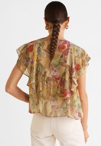 Przezroczysta bluzka w kwiaty z falbanistymi rękawami, w stonowanych kolorach zieleni, pomarańczy i różu na jasnym tle. Widoczne są białe spodnie.