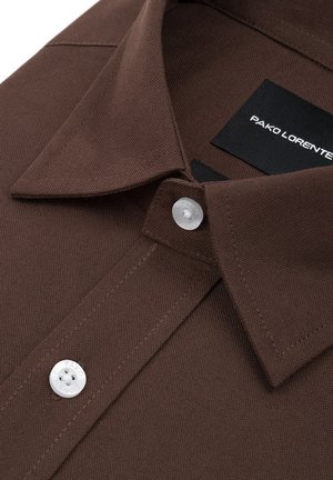 Zbliżenie na brązową koszulę z kołnierzykiem, białymi guzikami oraz czarną metką z napisem "PAKO LORENTE" wewnątrz kołnierza.