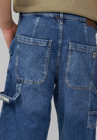 Pantalones cargo de mezclilla azul con un lavado desgastado, que presentan dos grandes bolsillos traseros y una etiqueta de marca en la cintura. Tejido texturizado y duradero.