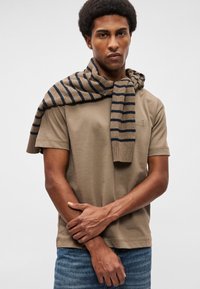 Barna, rövid ujjú póló, texturált logóval, bézs és tengerészkék csíkos pulóverrel a vállon. Farmernadrág.