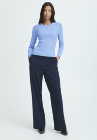 Una donna in piedi con le mani nelle tasche, che indossa un maglione a coste azzurro chiaro, pantaloni neri larghi e scarpe nere con punta su sfondo bianco.