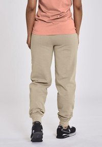 Beige Sweatpants mit einer weichen Textur, schmal zulaufenden Beinen und gerippten Bündchen, kombiniert mit schwarzen Turnschuhen. Das Oberteil ist ein schlichtes Pfirsichfarben.