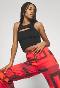 Top cropped preto com textura canelada e um recorte frontal, combinado com jeans soltos vermelhos com padrões geométricos castanhos e três botões decorativos.