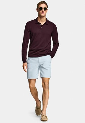 Uomo che cammina indossando occhiali da sole rotondi, polo a maniche lunghe viola scuro, pantaloncini azzurri e scarpe da barca beige su uno sfondo semplice.