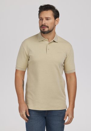 Mężczyzna z krótkimi ciemnymi włosami i brodą, ubrany w beżową koszulkę polo z krótkim rękawem i niebieskie dżinsy, stojący na gładkim jasnym tle.