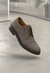 Καφέ δερμάτινο παπούτσι με κορδόνια και στρογγυλή μύτη, μαύρη αντίθετη σόλα και διακριτές ραφές. Το φόντο διαθέτει μινιμαλιστικό σχέδιο.