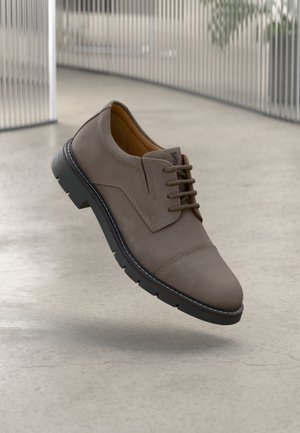 Zapato de cuero marrón con cordones y punta redondeada, con suela negra en contraste y detalles de costura sutiles. El fondo presenta un diseño minimalista.