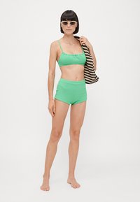 Set di bikini verde a coste composto da un top corto con spalline regolabili e pantaloni a vita alta. La modella tiene una borsa a righe.