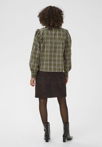 Blouse à carreaux verts avec manches bouffantes, associée à une jupe en daim marron foncé. L'ensemble présente une texture lisse et une coupe ajustée.