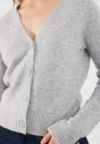Gaišpelēks kardigans, kas izgatavots no mīkstas auduma, ar dziļu V-veida kakla izgriezumu, ribotu apakšmalu un piedurknēm, kā arī pieciem kontrastējošiem pogām priekšpusē.