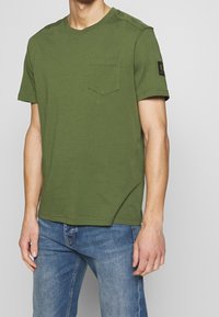 Man i grön t-shirt med korta ärmar, bröstfickan och ett märke på vänster ärm, tillsammans med blå jeans mot en enkel bakgrund.