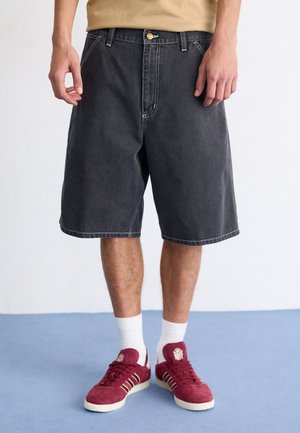 Personne portant un short en jean noir jusqu'aux genoux, des chaussettes blanches et des baskets rouges, debout sur un sol bleu.