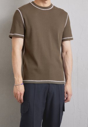 Camiseta marrón de canalé con mangas cortas, con costuras blancas en el cuello y los puños. Llevada con pantalones oscuros.