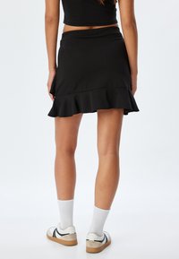 Zwarte uitlopende rok met een rucheszoom, gemaakt van zachte stof. Draagt met witte sokken en casual schoenen, wat een getailleerde silhouette toont.