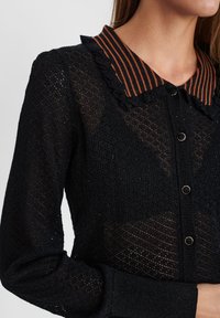 Musta neulottu neuletakki, jossa on raidallinen oranssi-musta röyhelökaulus, napillinen suljenta ja teksturoitu kuvio koko takissa.