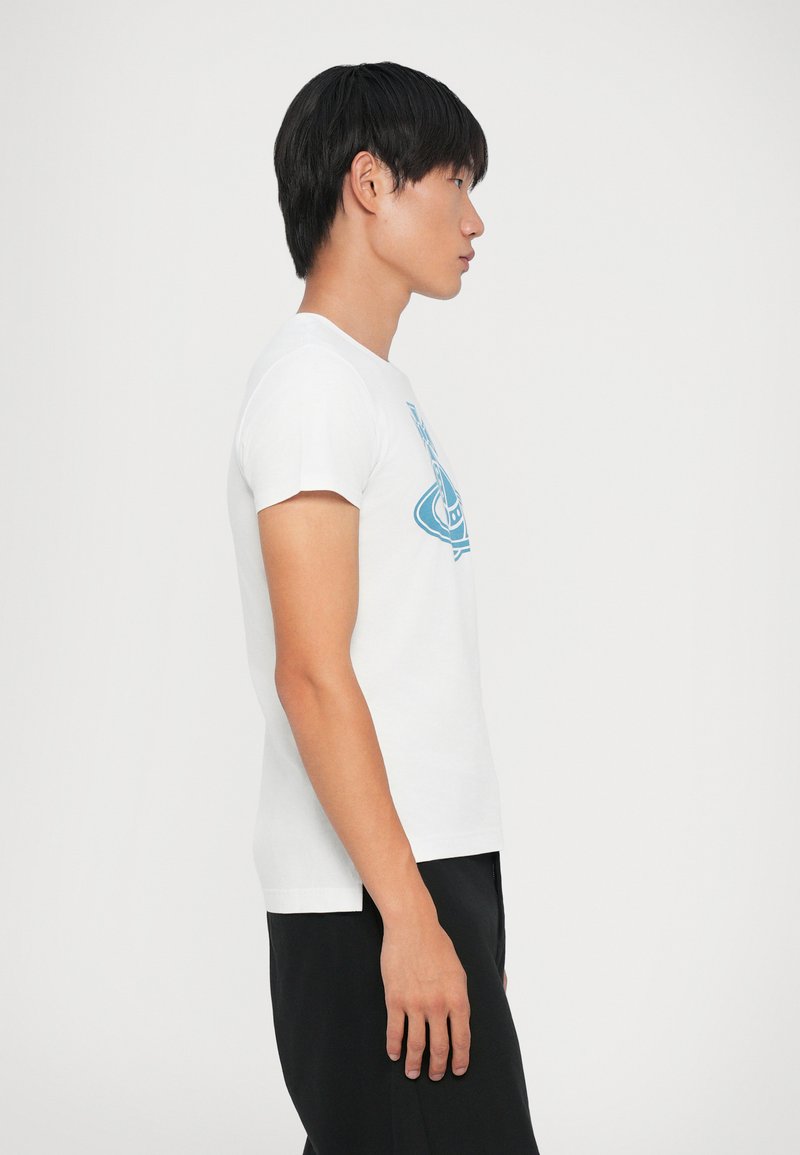 T-shirt en coton blanc à manches courtes et col rond, avec un design graphique bleu clair. Le mannequin porte un pantalon noir.