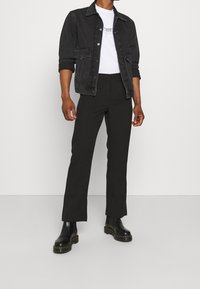 Mężczyzna w czarnej dżinsowej kurtce z podwiniętymi rękawami, białej koszulce z nadrukiem, czarnych spodniach o prostych nogawkach i czarnych skórzanych butach na tle białej ściany.