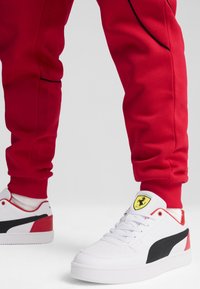 Person som bär röda byxor och vita Puma-sneakers med svart rand och gult Ferrari-logotyp på tungan, stående på vit golv.