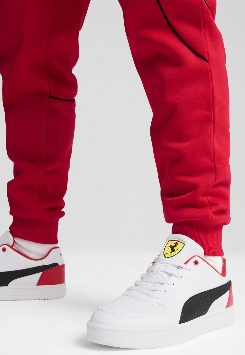 Person som bär röda byxor och vita Puma-sneakers med svart rand och gult Ferrari-logotyp på tungan, stående på vit golv.