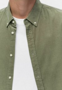 Vaaleanvihreä samettipaitapusero napituksella ja kauluksella, yhdistetty valkoiseen crewneck-allepaitaan, joka näkyy paidan alta.