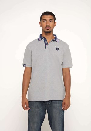Polo shirt gris à manches courtes avec un col marine arborant des rayures rouges et blanches, un patch avec logo et un logo sur le côté de la manche. Texture lisse.