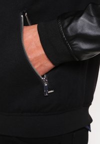 Sudadera negra con un bolsillo con cremallera. Presenta una manga de cuero texturizado, puños acanalados y un cuerpo de tela lisa. Los detalles de la cremallera son metálicos.