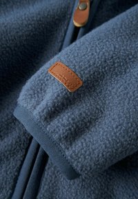 Niebieska rękawiczka polarowa z widoczną brązową skórzaną naszywką logo i uchwytem suwaka, prezentująca miękką teksturowaną tkaninę oraz detale szwów.