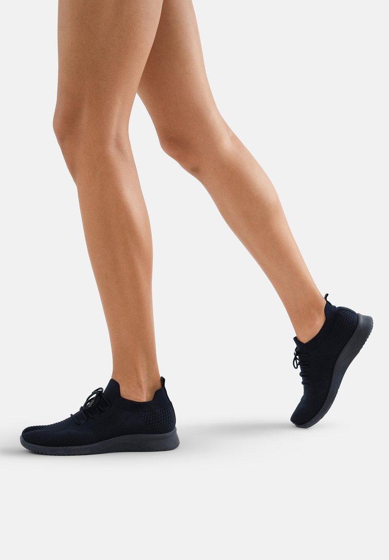 Chaussures de sport noires avec un dessus en tricot texturé, semelle légère et design à lacets, positionnées à un angle léger en marche.