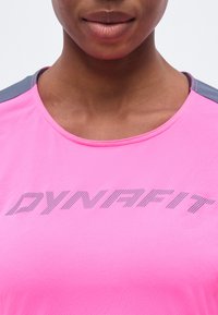 Κοντινό ενός ατόμου που φοράει φανέλα αθλητική σε φωτεινό ροζ χρώμα με γκρι λεπτομέρειες και την επιγραφή "DYNAFIT" στο στήθος.
