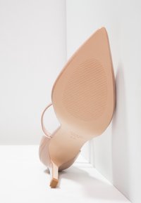 Nudové lakované boty na vysokém podpatku s špičatou špičkou a tenkým podpatkem. Podešev má texturovaný vzor pro lepší přilnavost.