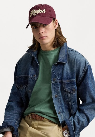 Denim jacket in dark blue, green crew neck t-shirt, maroon cap with white logo, brown braided belt, beige pants, and wristwatch.