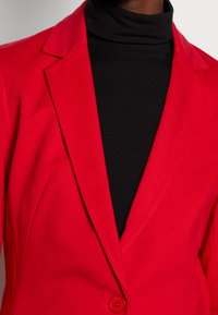 Czerwony żakiet o gładkiej teksturze, z wyłogi w szpic i jednolitym zapięciem na guzik, noszony z czarnym golfem pod spodem.