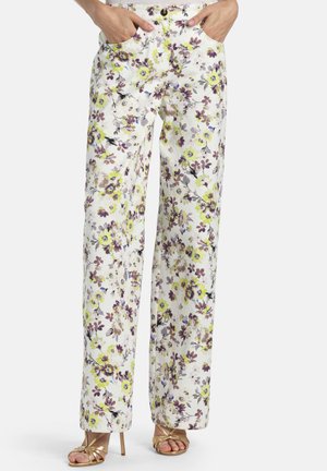 Biele nohavice s širokými nohavicami s kvetinovou potlačou v žltej, fialovej a sivej farbe, nosené so zlatými sandálmi na vysokom opätku s otvorenou špičkou a rukami v kapsách.