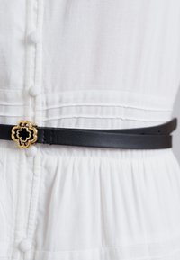 Cinturón de cuero negro con una hebilla floral dorada, usado sobre una tela blanca texturizada. Presenta una textura suave y un diseño minimalista.