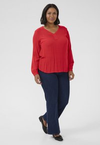 Blusa rossa a maniche lunghe con scollatura a V, dettagli plissettati e texture morbida abbinata a jeans svasati blu scuro e scarpe nere basse.