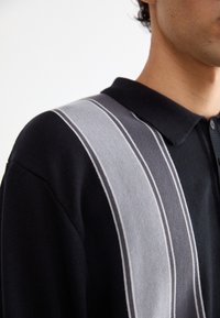 Schwarzes Poloshirt mit grauen und hellgrauen Streifen, mit einem kragenartigen Ausschnitt und feiner Strickstruktur. Nahaufnahme des Oberteils.