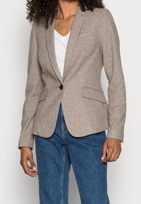 Blazer texture marrone chiaro con un singolo bottone rotondo, due tasche frontali e un taglio sartoriale, indossato sopra una maglietta bianca a V e jeans blu.