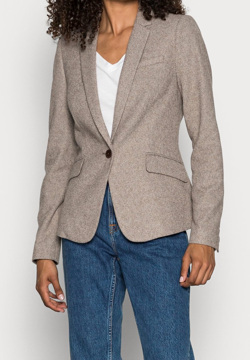 Blazer texture marrone chiaro con un singolo bottone rotondo, due tasche frontali e un taglio sartoriale, indossato sopra una maglietta bianca a V e jeans blu.