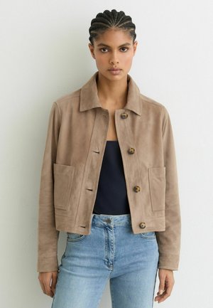 Nuori nainen, jolla on letitetty tukka ja beige mokkatakki isoilla taskuilla, musta paita ja vaaleansiniset farkut, seisoo valkoista seinää vasten.