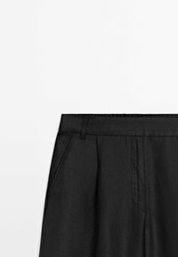 Pantalon noir taille haute avec passants de ceinture, poches latérales et une couture visible sur la braguette devant, sur un fond clair uni.