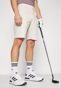 Beiga golfshorts med rak front, bärs med vita och marinblå sportskor, svartvita randiga strumpor och håller en golfklubba.