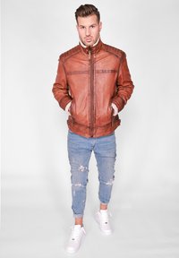 Bruine leren jas met een hoge kraag, genuanceerde schouderaccenten en twee zijzakken, in combinatie met lichtblauwe ripped jeans en witte sneakers.