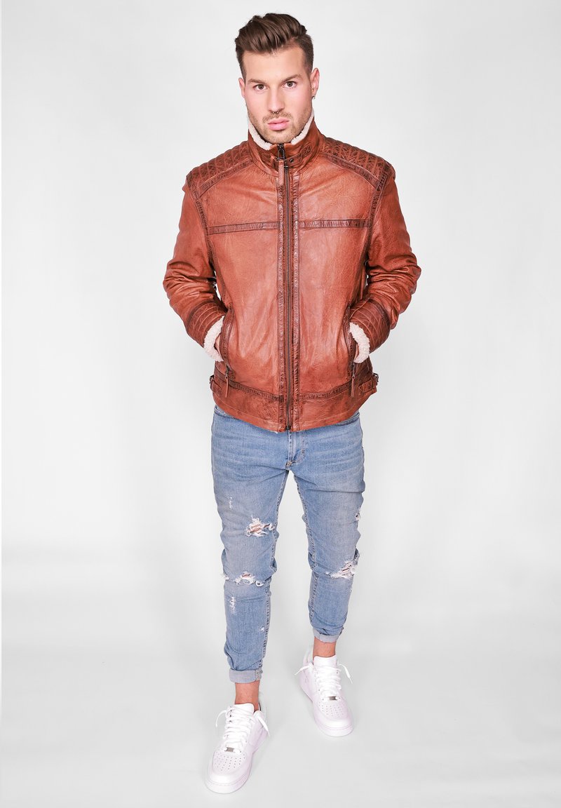 Bruine leren jas met een hoge kraag, genuanceerde schouderaccenten en twee zijzakken, in combinatie met lichtblauwe ripped jeans en witte sneakers.